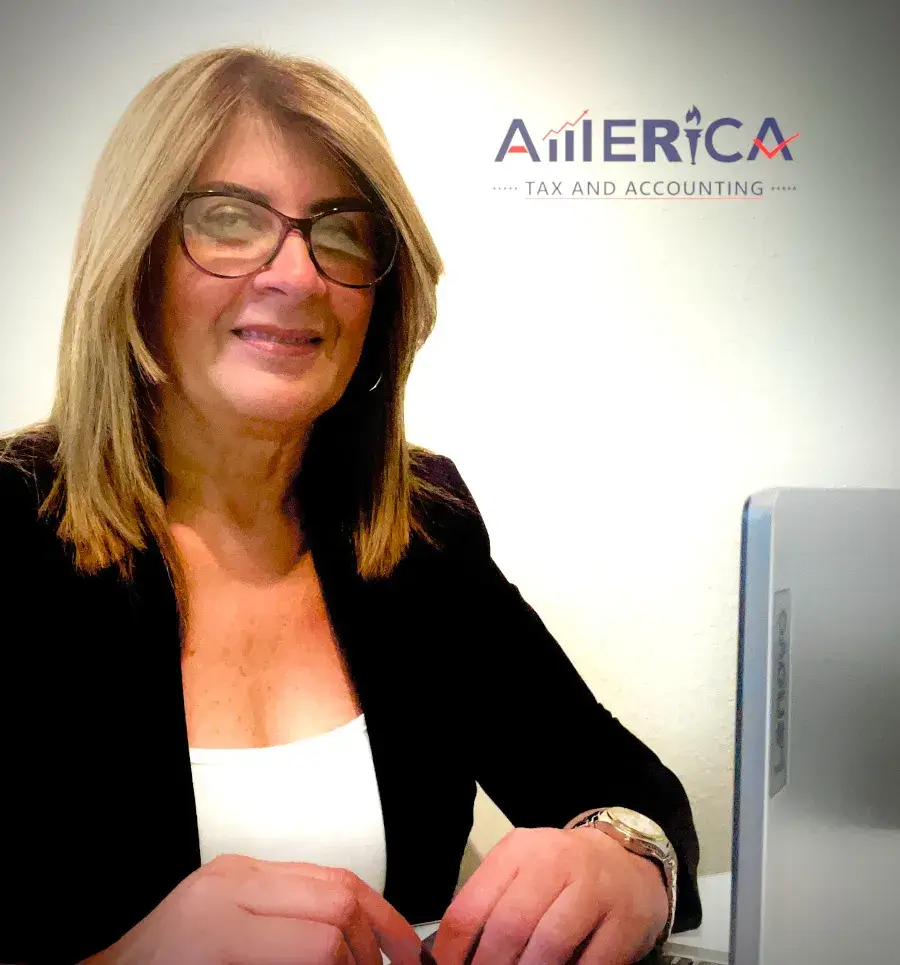 Image of bookkeeper, accountant, and tax preparer Claudia Baez sitting at a desk in front of a white backdrop with her computer. Claudia is wearing a black business suit and white shirt. There is also an America Tax & Accounting logo on the white background.