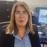 Image of woman speaking into camera. There's an IRS logo on the foreground. The woman is wearing a black business suit and white collar shirt. There's a computer with the America Tax & Accounting, Corp. Logo in the background.